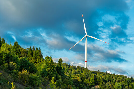Modern wind turbine generating renewable energy against blue sky with white clouds above lush green forest landscape in countryside - Powered by Adobe