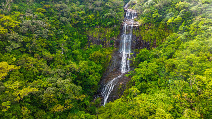 Aerial view of beautiful tiered waterfall cascading down rocky cliff face surrounded by dense lush green tropical rainforest canopy