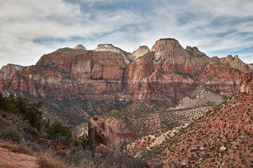 A wide view of tall sandstone cliffs showing warm bands of light and dark rock. The layered walls rise above a dry foreground with low brush, natural texture in the stone.
