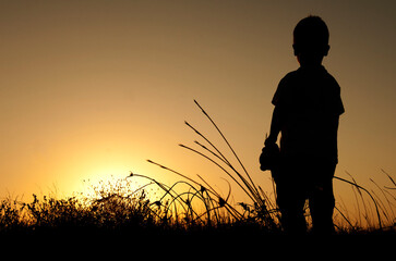 Silhouette of a young child standing in tall grass against a vibrant sunset, capturing a moment of wonder and exploration in nature's beauty and tranquility