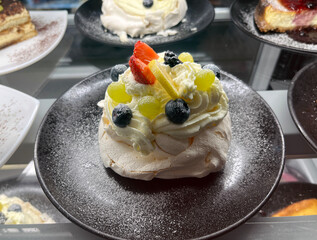 Close-up of a pavlova dessert topped with whipped cream, strawberries, blueberries, and grapes, served on a black plate with powdered sugar garnish.