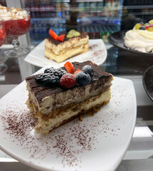 Close-up of a slice of tiramisu on a white plate, topped with fresh berries and dusted with cocoa powder, displayed in a dessert showcase.