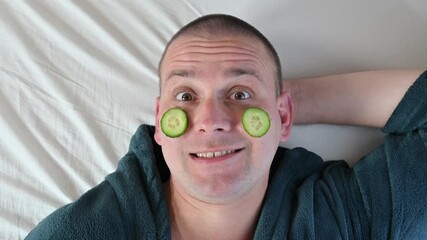 Man applies a cucumber face mask, relaxing in a bathrobe during skincare routine, close-up wellness concept.