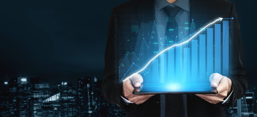 A professional businessman stands in a modern city at night, holding a tablet displaying digital graphs and data that illustrate business growth and success. Impute