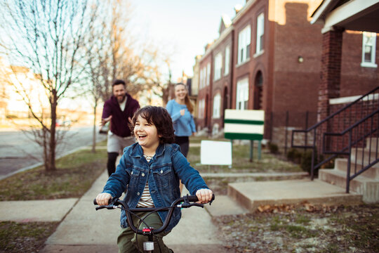 Happy child rides bike with adult parents on neighborhood sidewalk