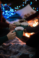 Cozy winter gathering with friends holding warm mugs near fire and festive lights concept of holiday decor and christmas warmth