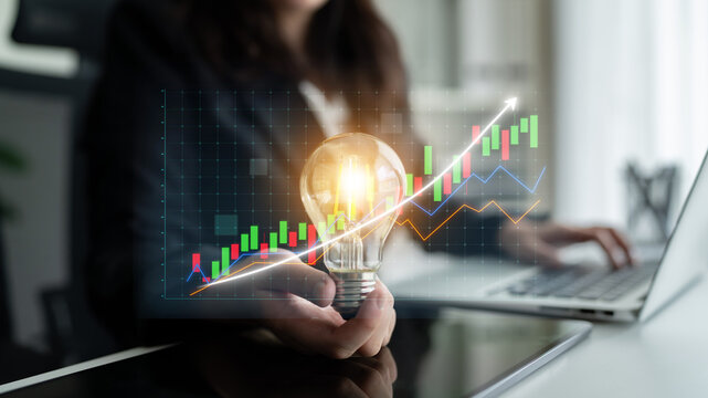 A business professional holds a light bulb, representing innovation, while viewing digital financial graphs on a laptop in a modern office environment. Impute - Powered by Adobe