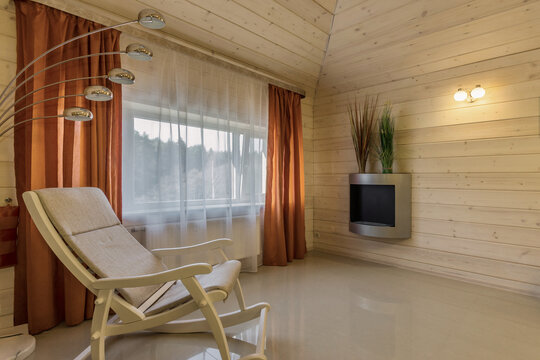 A comfortable cabin interior showcases a sleek chair and large window with orange curtains. The space is inviting with light wood walls and a modern fireplace. Natural light fills the room
