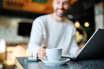 Man smiling, holding coffee while working on a laptop in a cafe