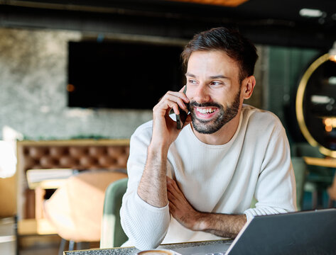 Man smiling, holding coffee while working on a laptop and using a mobile phone in a cafe