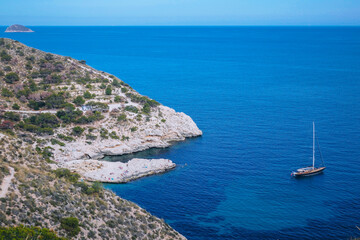 A serene coastal scene featuring a rocky shoreline and a calm blue sea. A sailboat is anchored near...
