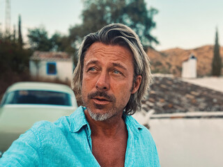Tanned middle aged man with half long blond hair in blue shirt by classic car at white spanish house in the countryside. Golden hour in Malaga, Andalusia, Spain.