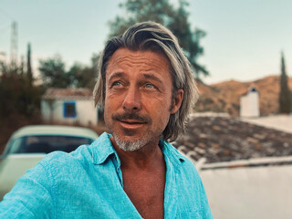 Tanned middle aged man with half long blond hair in blue shirt by classic car at white spanish house in the countryside. Golden hour in Malaga, Andalusia, Spain.