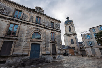 Obraz premium A low-angle view of a historic square in La Rochelle featuring a weathered stone facade and a tall belfry clock tower under an overcast sky, showcasing classic French architecture and cobblestone