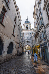 A street view in La Rochelle looking towards the iconic Grosse Horloge clock tower gate. Wet cobblestones and historic stone buildings frame the medieval archway under an overcast sky.