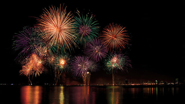 Spectacular colorful fireworks light up the dark night sky, reflecting brilliantly on calm water. A distant city skyline adds festive ambiance. - Powered by Adobe