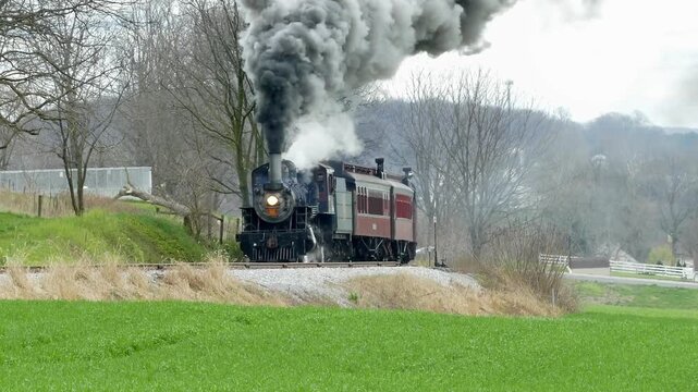 A vintage steam train chugs along a rural track surrounded by green fields and bare trees. The sound of the engine echoes as it fills the air with smoke.