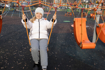 A woman is swinging on a round baby swing on a playground. She is wearing a white jacket and a knitted hat.
