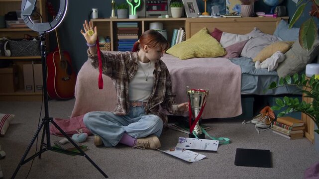 Long shot of gen Z Caucasian girl demonstrating medals, certificates and trophies while vlogging on smartphone at home