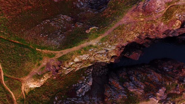 Grassy ridge above narrow coastal fissure. A winding path follows a green ridge beside a steep rocky split overlooking the sea.