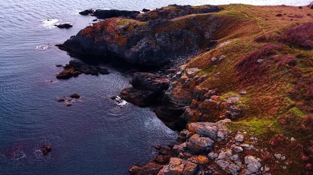 Sunlit cliffs rising from dark waters. Warm light highlights rugged rock faces meeting calm deep blue sea along the shore.