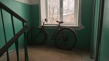 Vintage black bicycle stands near a window on a gloomy apartment building stairwell landing, framed by the green stair railing and turquoise walls