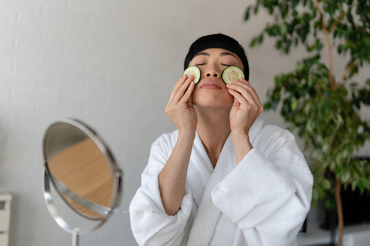 Asian woman in a bathrobe applying fresh cucumber slices to her face while doing a relaxing home skincare routine at a table with beauty products
