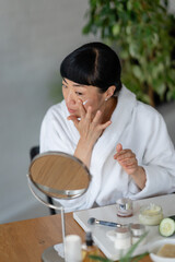 Asian woman in a white robe applying face cream while looking in a mirror; the mood is calm,...