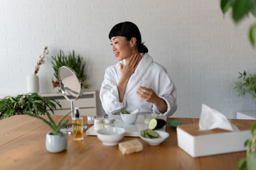 Asian woman in a bathrobe touching her neck while preparing skincare products during a calm home...