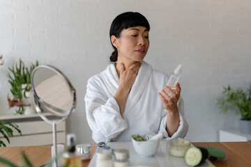 Asian woman in a bathrobe examining a skincare serum bottle while touching her neck during a calm...