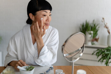 Asian woman in a white bathrobe applying face cream to her cheek, captured in a calm and focused skincare moment