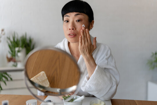 Asian woman in a white bathrobe applying face cream to her cheek, captured in a calm and focused skincare moment