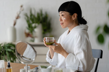 Woman in a white robe enjoying a warm drink during a relaxing self-care moment, sitting at a table with skincare products and a mirror