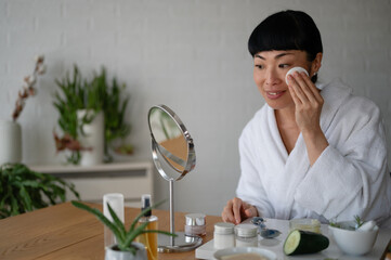Woman in a white bathrobe cleansing her face with a cotton pad during a home skincare routine, sitting at a table with beauty products and a mirror
