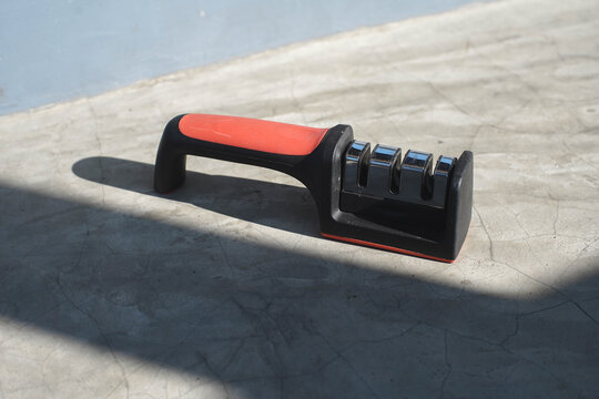 Modern kitchen knife sharpener with red and black handle on concrete surface, bathed in sunlight with strong shadows.