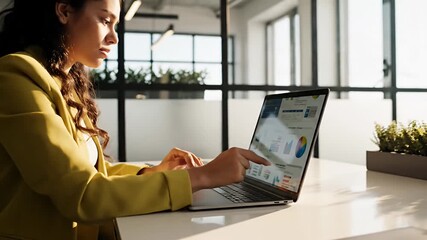 Young professional woman analyzing business data and marketing reports on a laptop in a modern office pointing at charts and graphs for strategic planning - Powered by Adobe