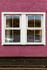 Fototapeta premium Glazed, double hung window reflecting the harbor with some boats, set in a purplish-pink painted, ancient fisherman's house. Portree-Skye-Scotland-091
