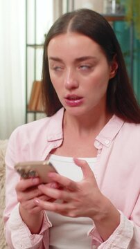 Young woman sitting on home sofa angrily types on smartphone chatting and waves hand in frustration at negative messages. Caucasian girl shows displeased look and posture reacting to words online.