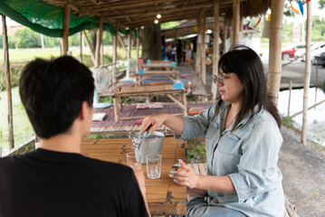 Two LGBTQ friends enjoying their lunch at an Isaan Thai restaurant. One is serving ice cubes into glasses while the other is seated, sharing a moment of connection in a cozy, vibrant atmosphere.