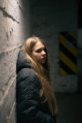 Moody urban portrait of a woman in a black puffer jacket, leaning against a white brick wall in dim industrial light.
