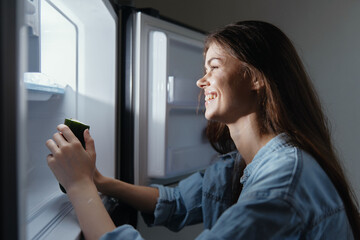 Young woman with long hair smiles while holding a green smoothie in front of an open refrigerator, representing healthy eating and lifestyle choices