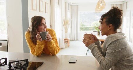 Diverse female friends raising mugs at kitchen island, sharing joke, laughing and sipping coffee