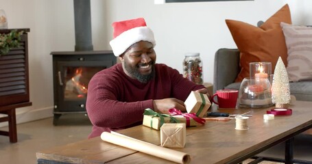 African American man in Santa hat lifting box at table tying ribbon bow for holiday gift - Powered by Adobe