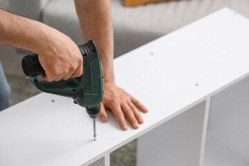 Young man with drill assembling shelf unit at home, closeup