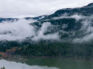 Fototapeta premium Misty Mountain Ridge Over Calm Lake in BC, Canada With Fog and Evergreen Forest