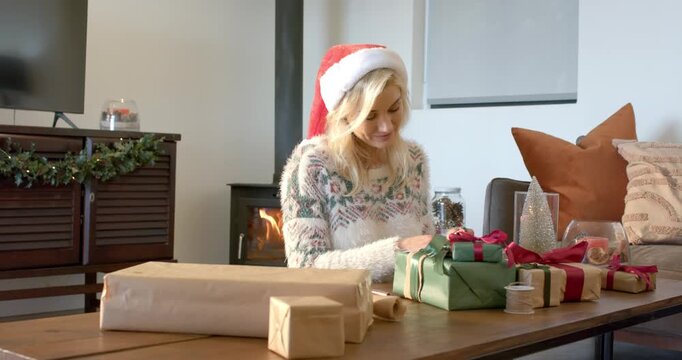 Woman wearing Santa hat wrapping gifts tying red ribbon at living room table by wood-burning stove