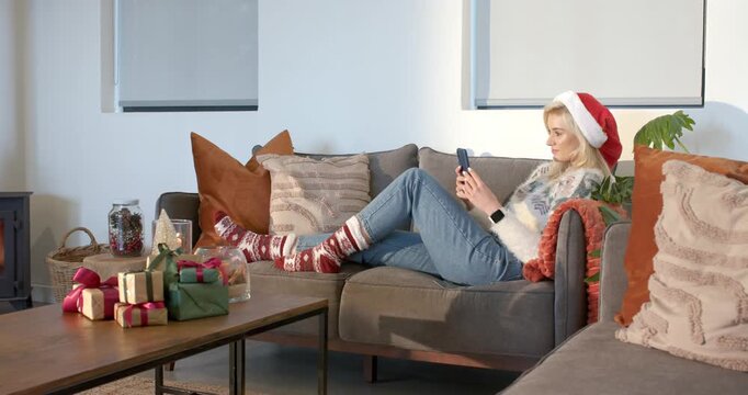 After settling on sofa, woman wearing Santa hat scrolling smartphone checking messages, copy space