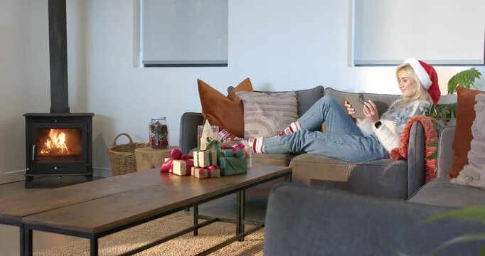 Woman wearing Xmas sweater lounging on sofa by stove scrolling phone, grabbing tablet, copy space