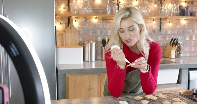 Woman in apron gripping icing bag piping icing onto cookies at kitchen counter explaining technique