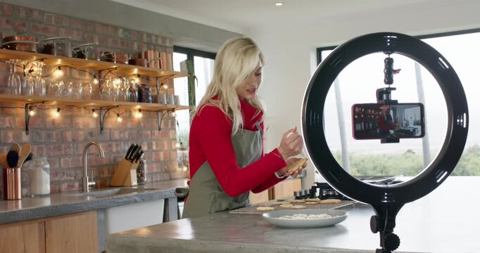 Adjusting smartphone starting recording, woman piping icing onto cookies for tutorial in kitchen